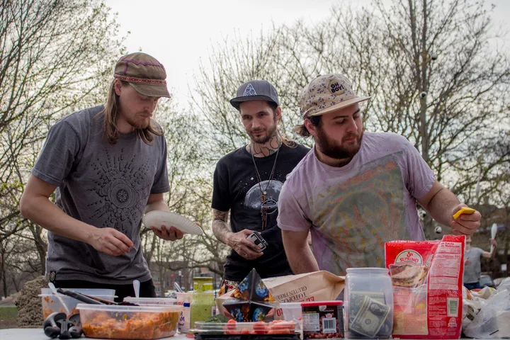 Three Taco Tuesday regulars preparing food at a crowded outdoor table in Palmer Square Park